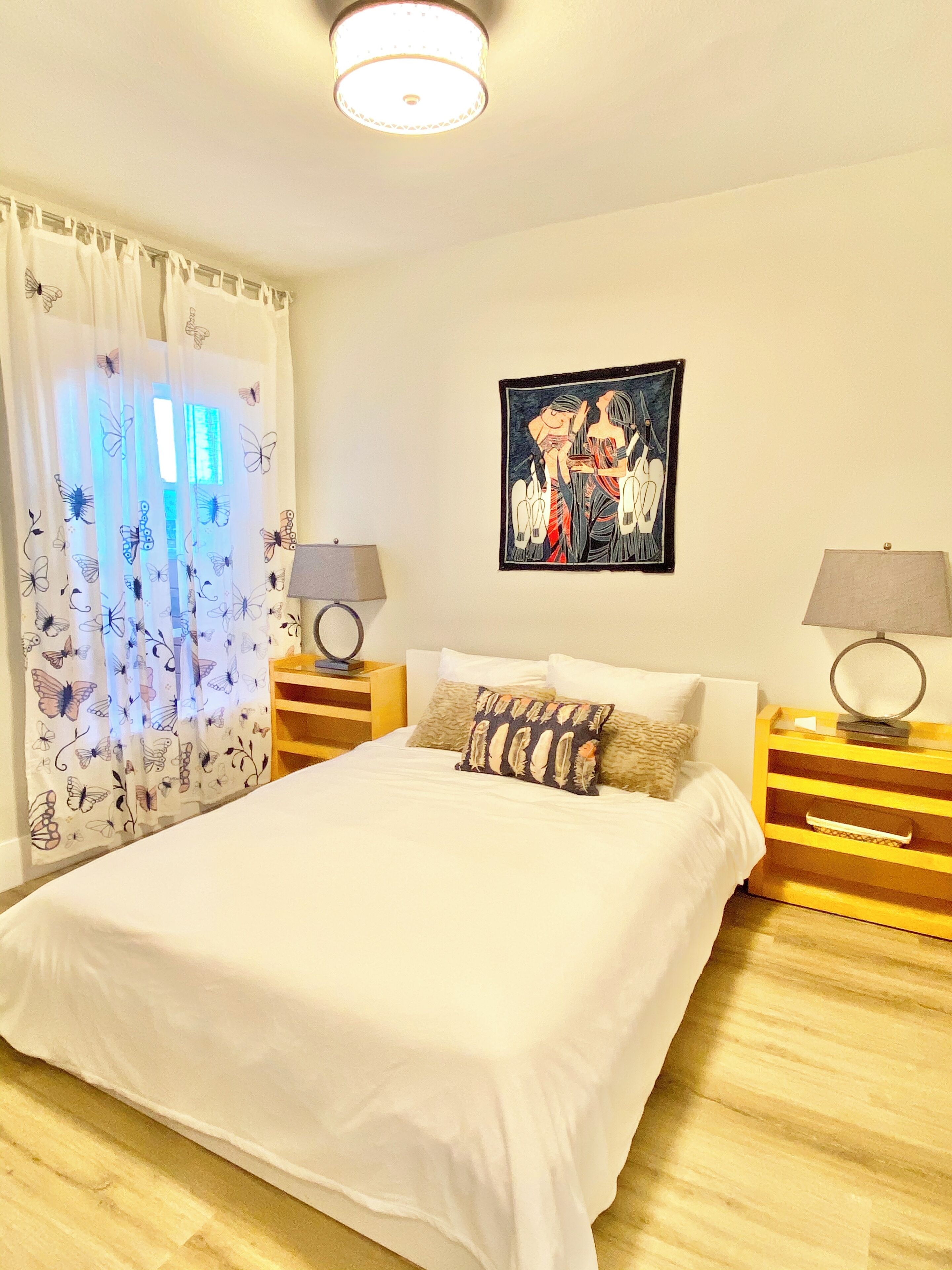 Une chambre à coucher avec un grand lit, deux tables de chevet avec des lampes, un tableau au mur et une fenêtre ornée de rideaux à motifs de papillons.
