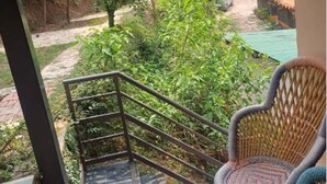 A balcony with a wicker chair, a metal railing, and a view of a green landscape with trees and a distant building. - PHS KASAULI HILLS (Krishangarh)