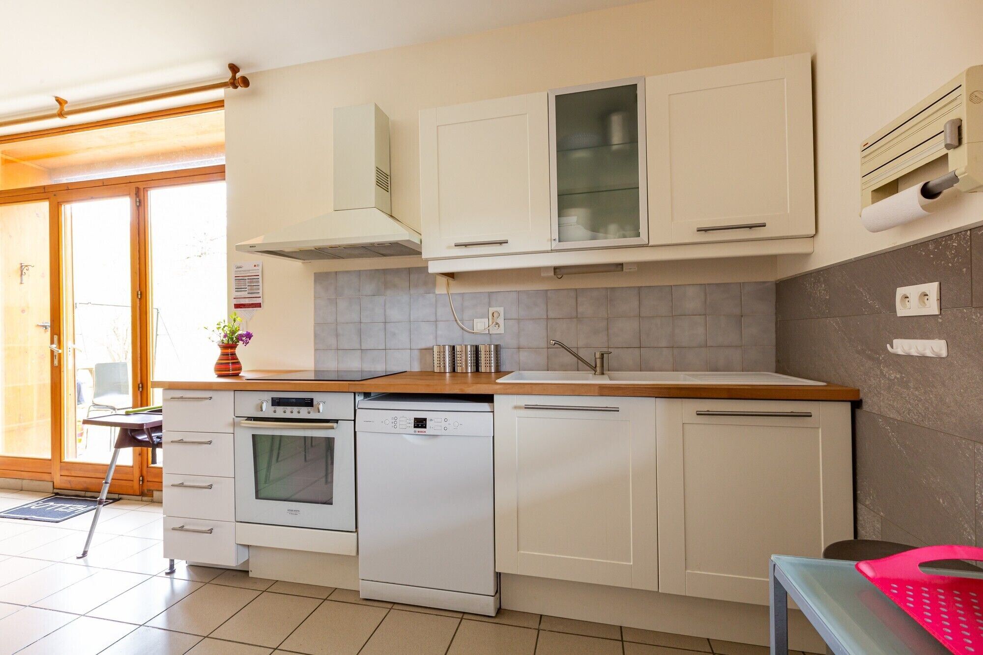 Una cucina moderna con mobili bianchi, forno integrato, lavastoviglie e una finestra con vista su un terrazza in legno.