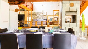 A dining area with a table set for four, surrounded by chairs, with a stone fireplace and bookshelves in the background.