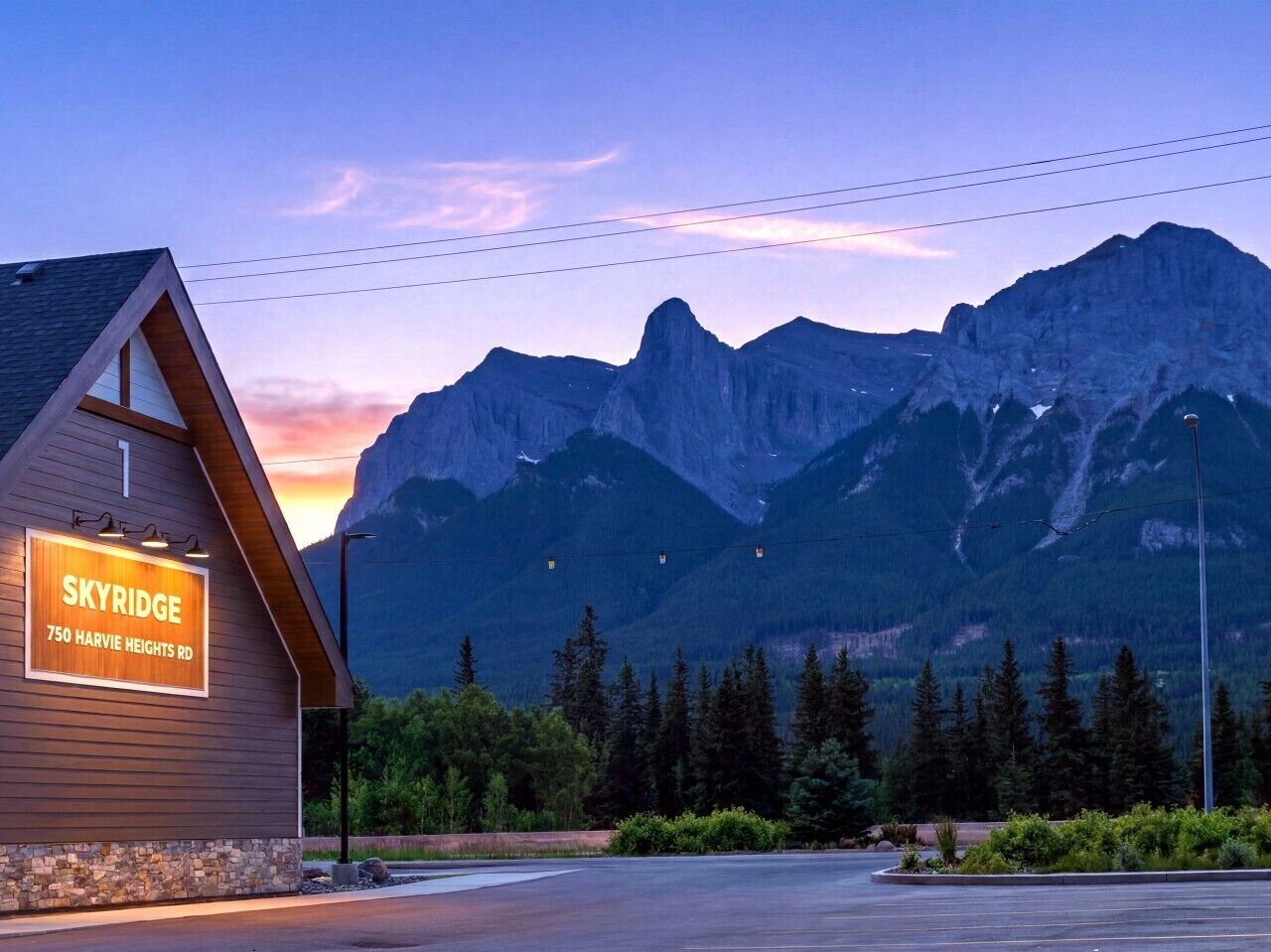 A building with a sign reading 'SKYRIDGE 750 HARVIE HEIGHTS RD' against a backdrop of mountains and a twilight sky.