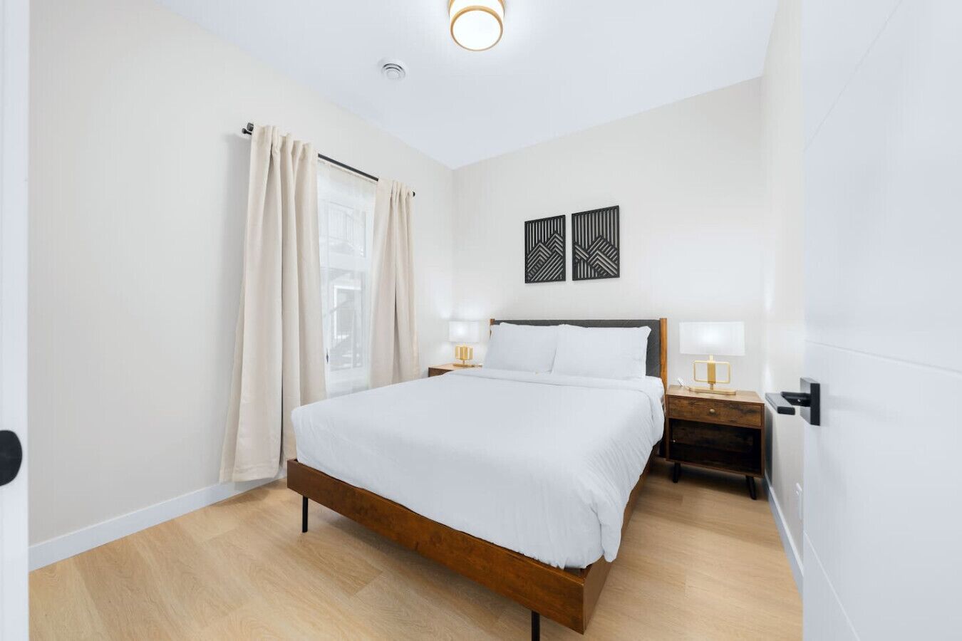 A modern bedroom with a wooden bed, a nightstand with a lamp, and a window with curtains.
