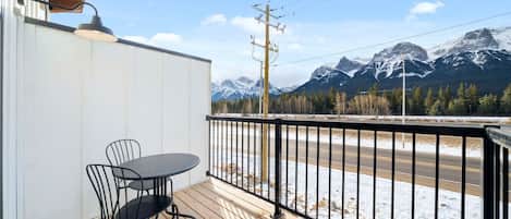 Eine Holzterrasse mit Tisch und Stühlen, einem schwarzen Geländer und Blick auf schneebedeckte Berge.