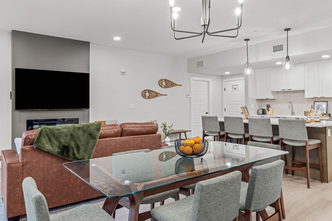 A modern living room with a glass dining table, grey upholstered chairs, a brown leather sofa, and a flat-screen TV mounted on the wall.