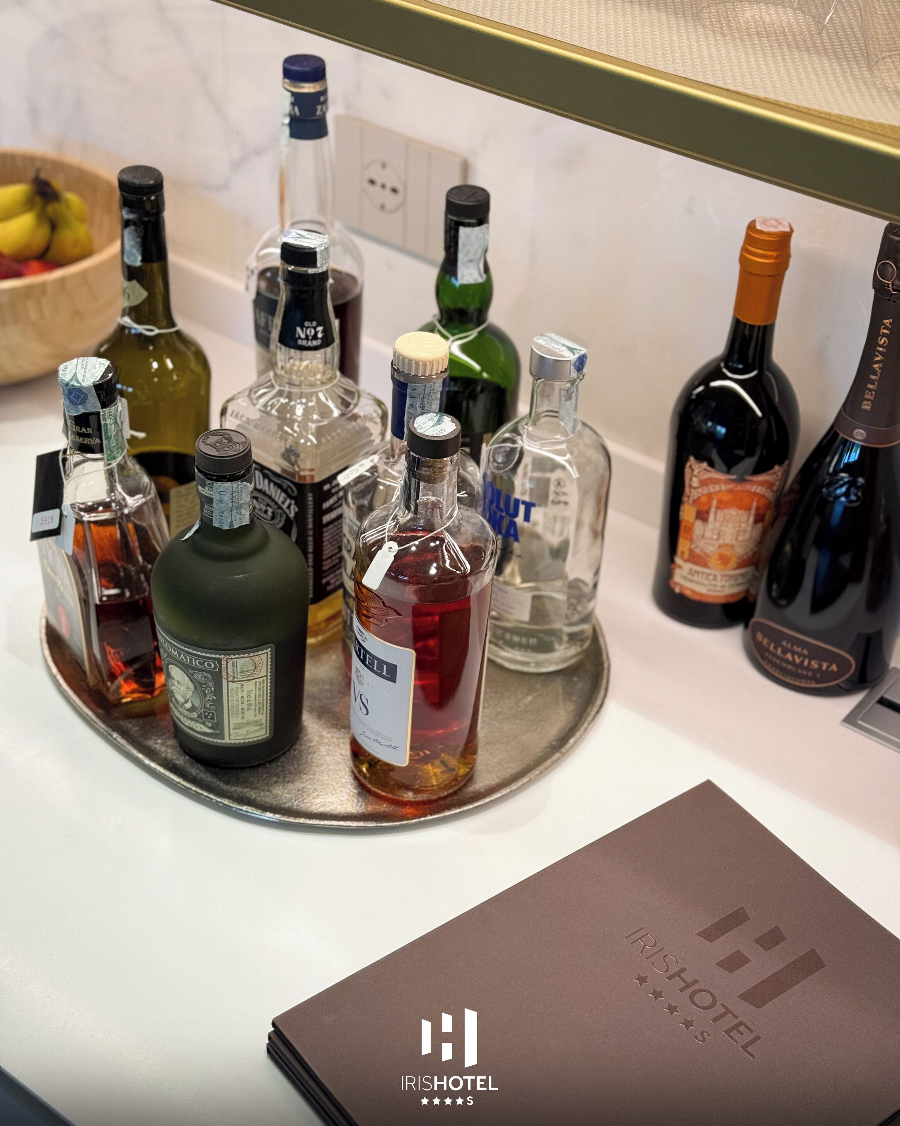 Una bandeja con varias botellas de licor, una carpeta marrón con el logotipo de IRISHHOTEL, y un frutero al fondo.
