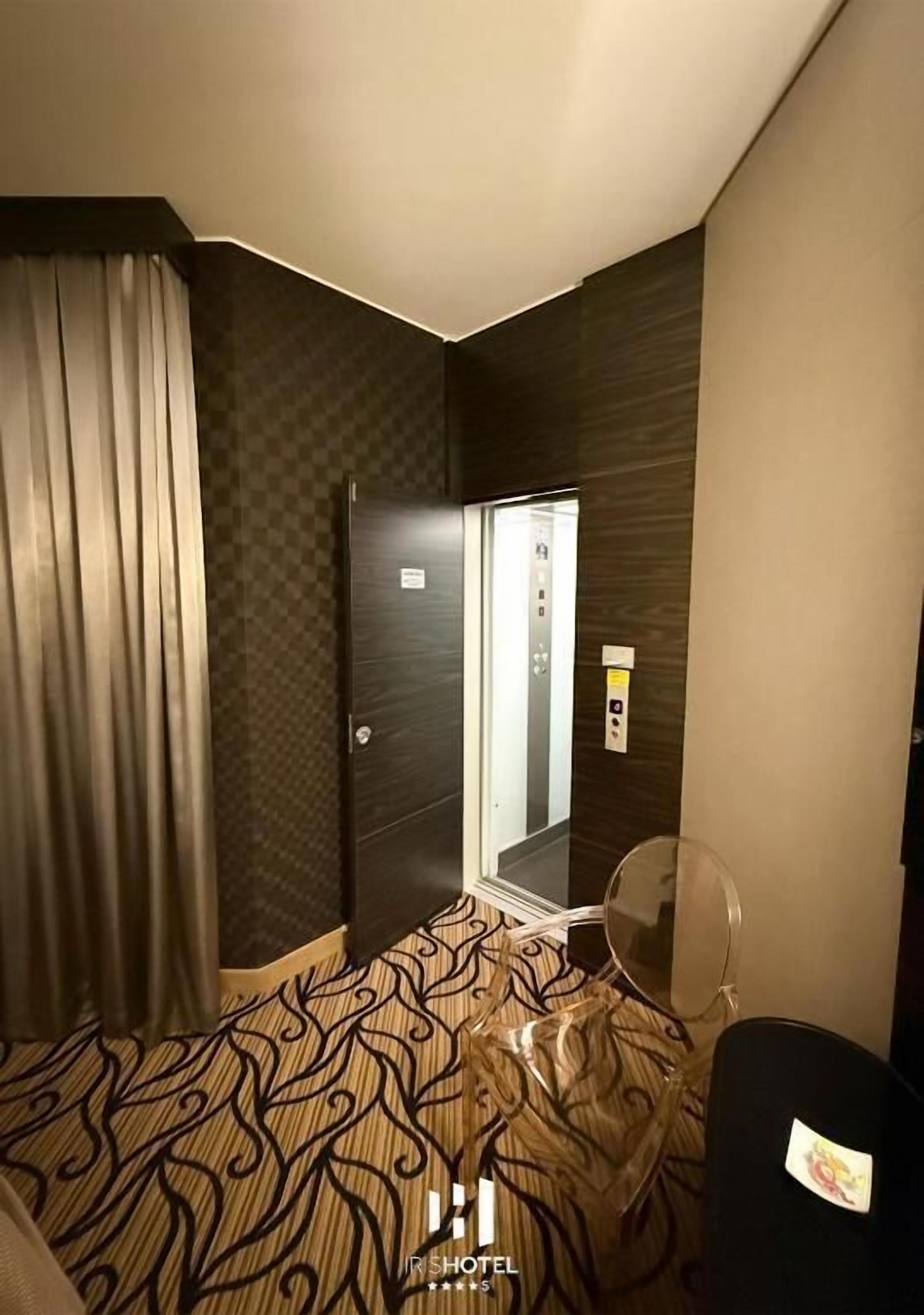 Quarto de hotel com uma cama, uma cadeira, uma mesinha e uma porta de elevador.