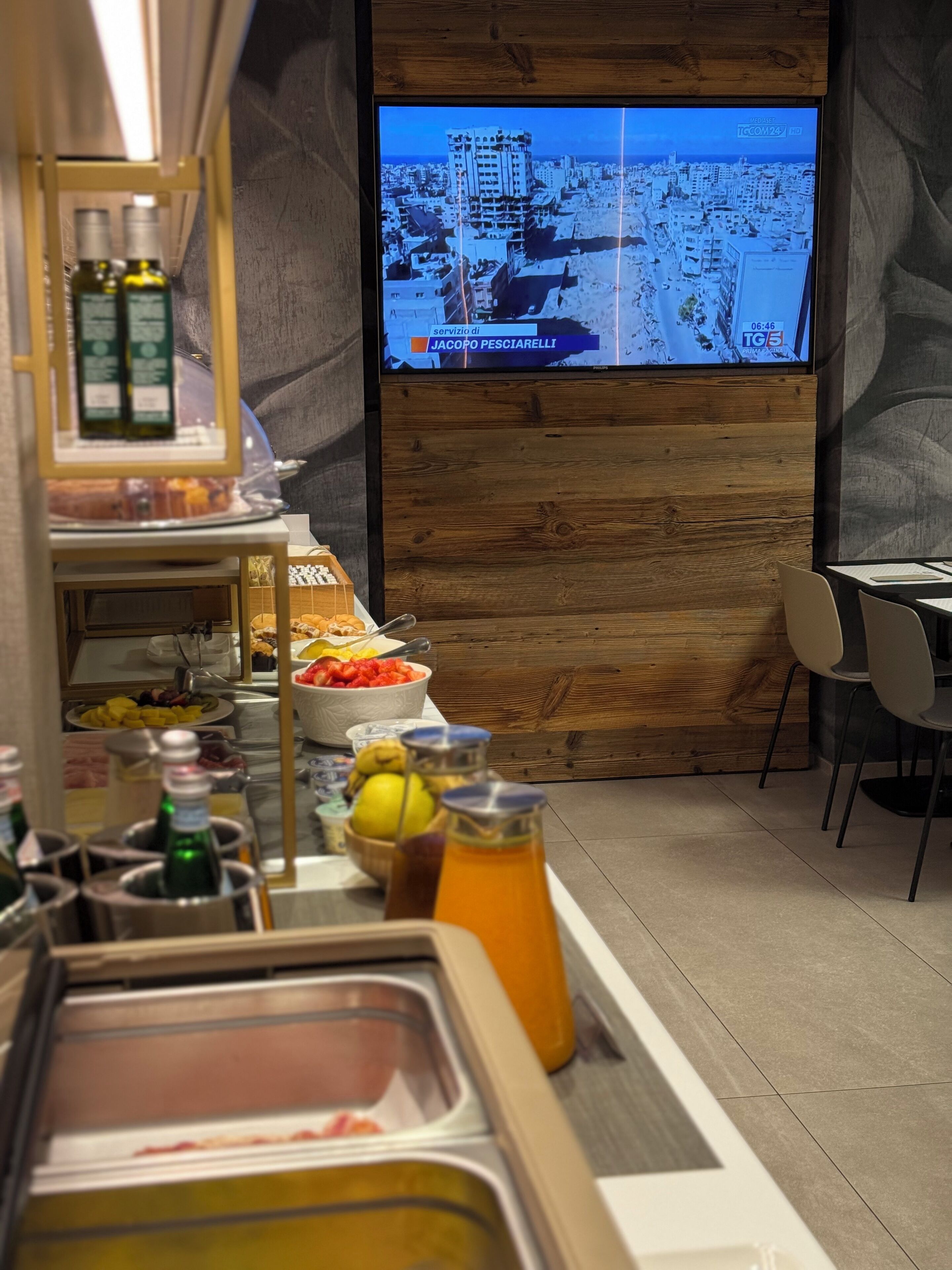 Un bufé de desayuno con diversos alimentos y bebidas, un televisor mostrando un noticiero y una pared de madera.
