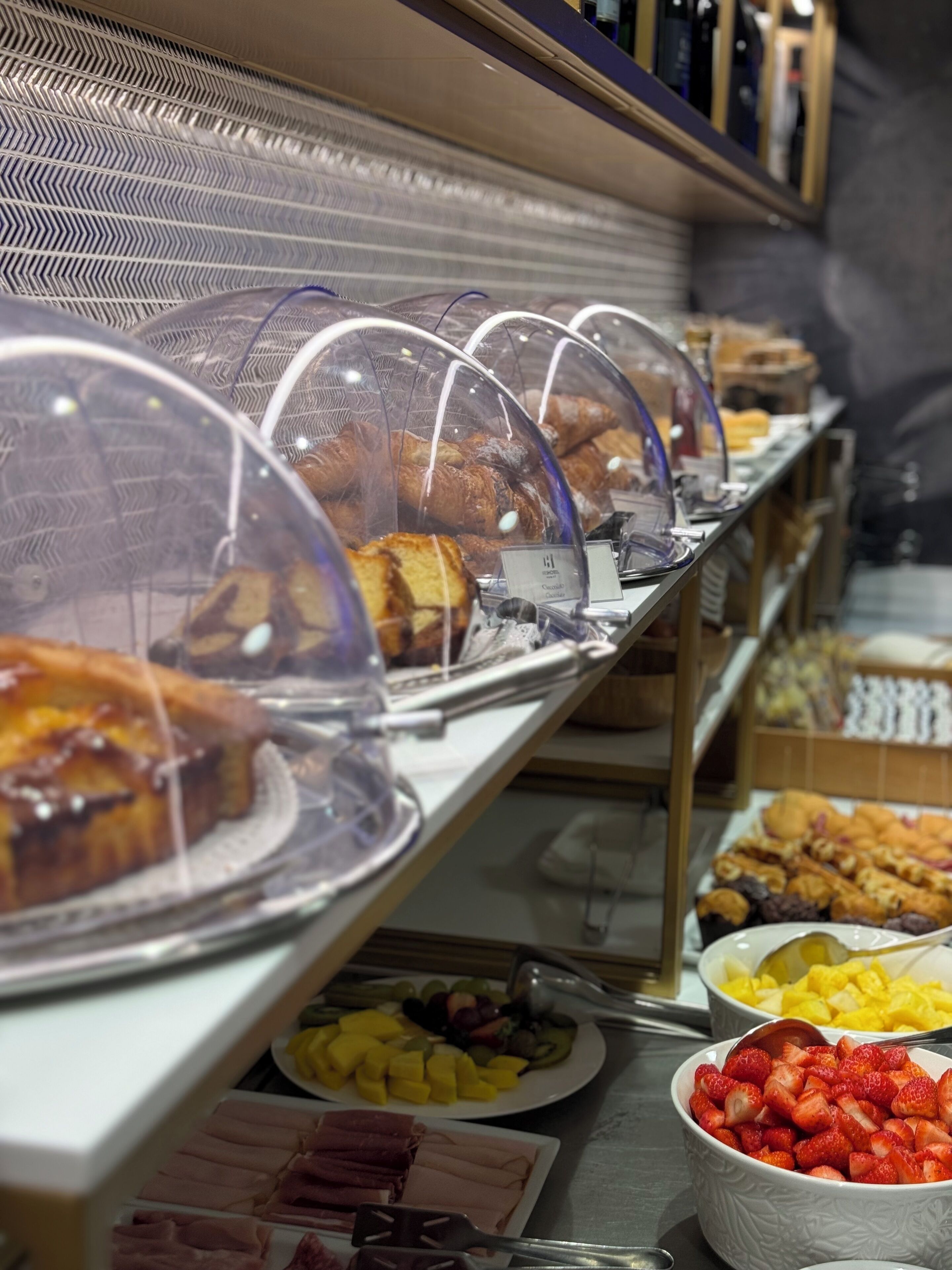A buffet with various food items under glass domes, including pastries, fruits, and meats.