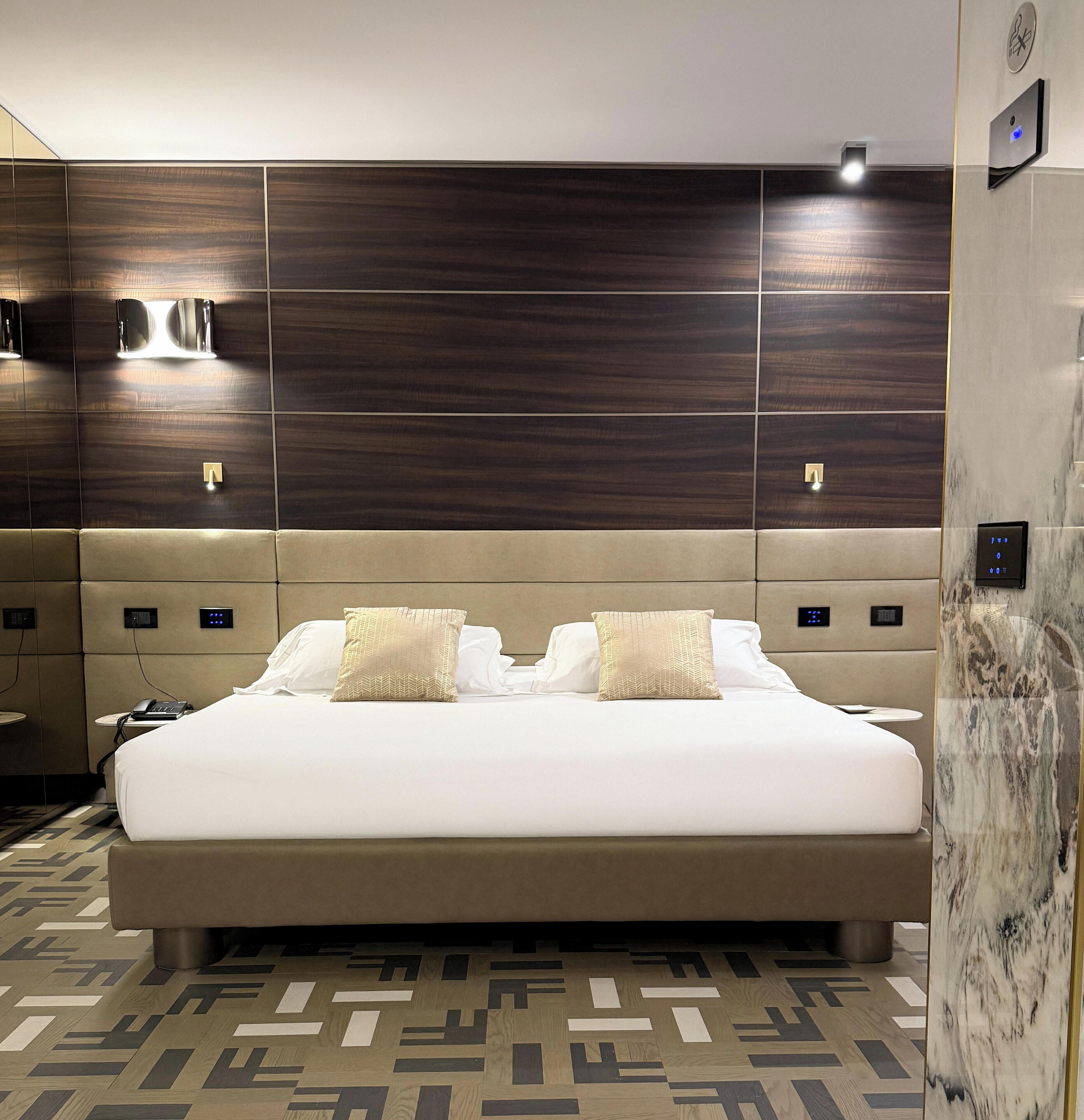 Habitación de hotel moderna con una cama grande, cabecera con iluminación integrada, mesita de noche con teléfono y suelo estampado.