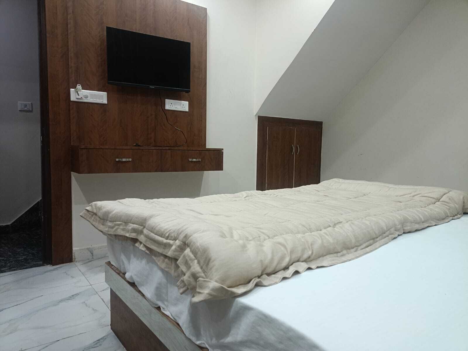A bedroom with a bed, a wooden wardrobe, a television mounted on the wall, and a small cabinet.
