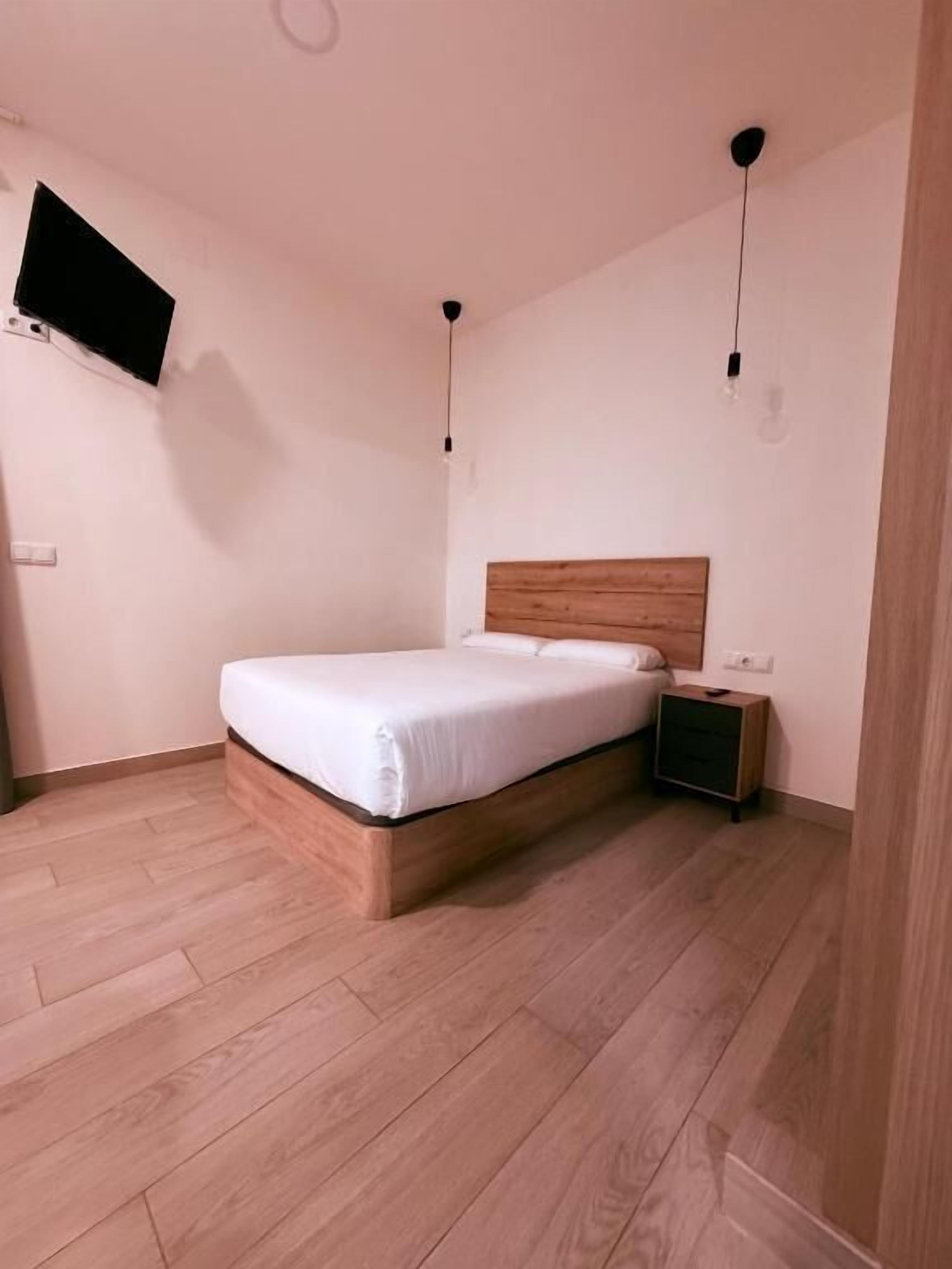 Un dormitorio con cama de madera, mesita de noche, televisor montado en la pared y piso de madera.