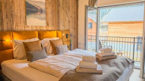 A bedroom with a wooden headboard, a large bed with white linens, a painting of a landscape, and a balcony with a view of a building. - Stylish chalet at lake Afritzer See with sauna (Afritz am See)