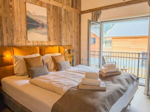 Quarto com cabeceira de madeira, cama grande com roupa de cama branca, pintura de paisagem e varanda com vista para um edifício.