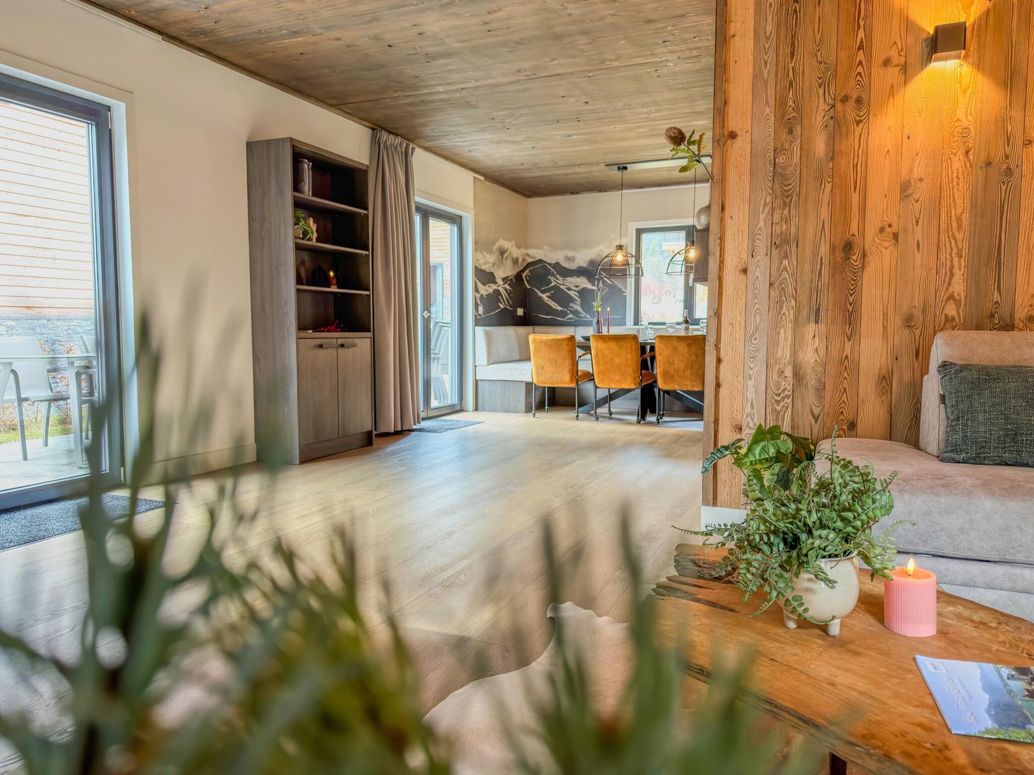 Un salon moderne avec un plafond en bois, un coin repas comprenant une table et des chaises, et un canapé agrémenté d’une plante et d’une bougie posées sur une table en bois.