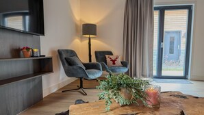 A modern living room with a wooden coffee table, two grey armchairs, a floor lamp, a TV mounted on the wall, and a sliding glass door leading outside. - Premium chalet with sauna on an alpine lake (Afritz am See)