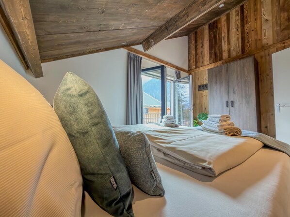 A bedroom with a bed, pillows, a wooden ceiling, a window with curtains, and a view of mountains.