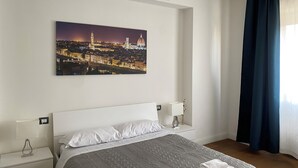 Ein Hotelzimmer mit einem Bett, einem Nachttisch, einer Lampe, einem Stadtbild und einem Fenster mit Vorhängen.