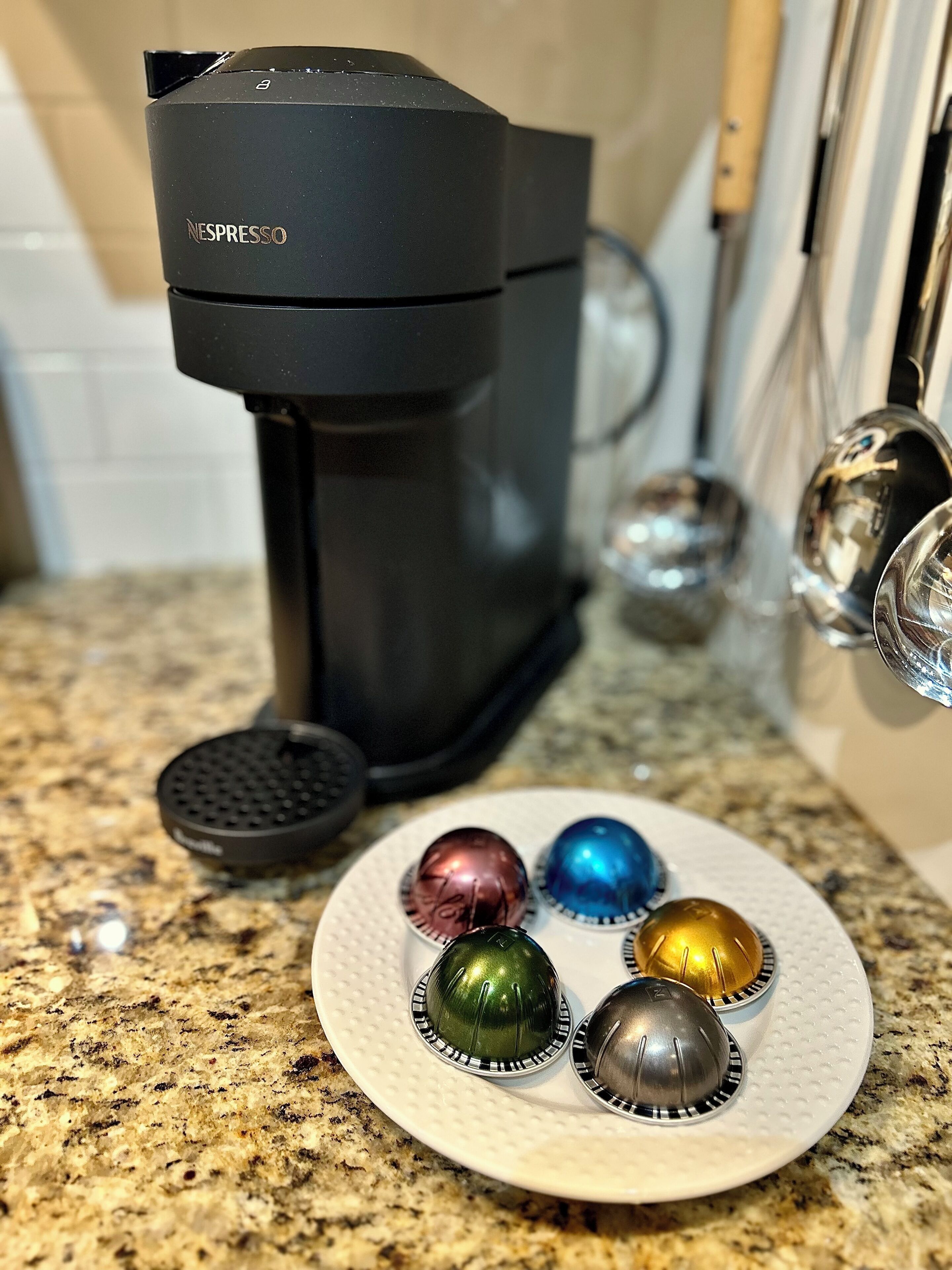 Une machine à café Nespresso posée sur un plan de travail de cuisine, avec une assiette remplie de capsules de café aux couleurs vives.