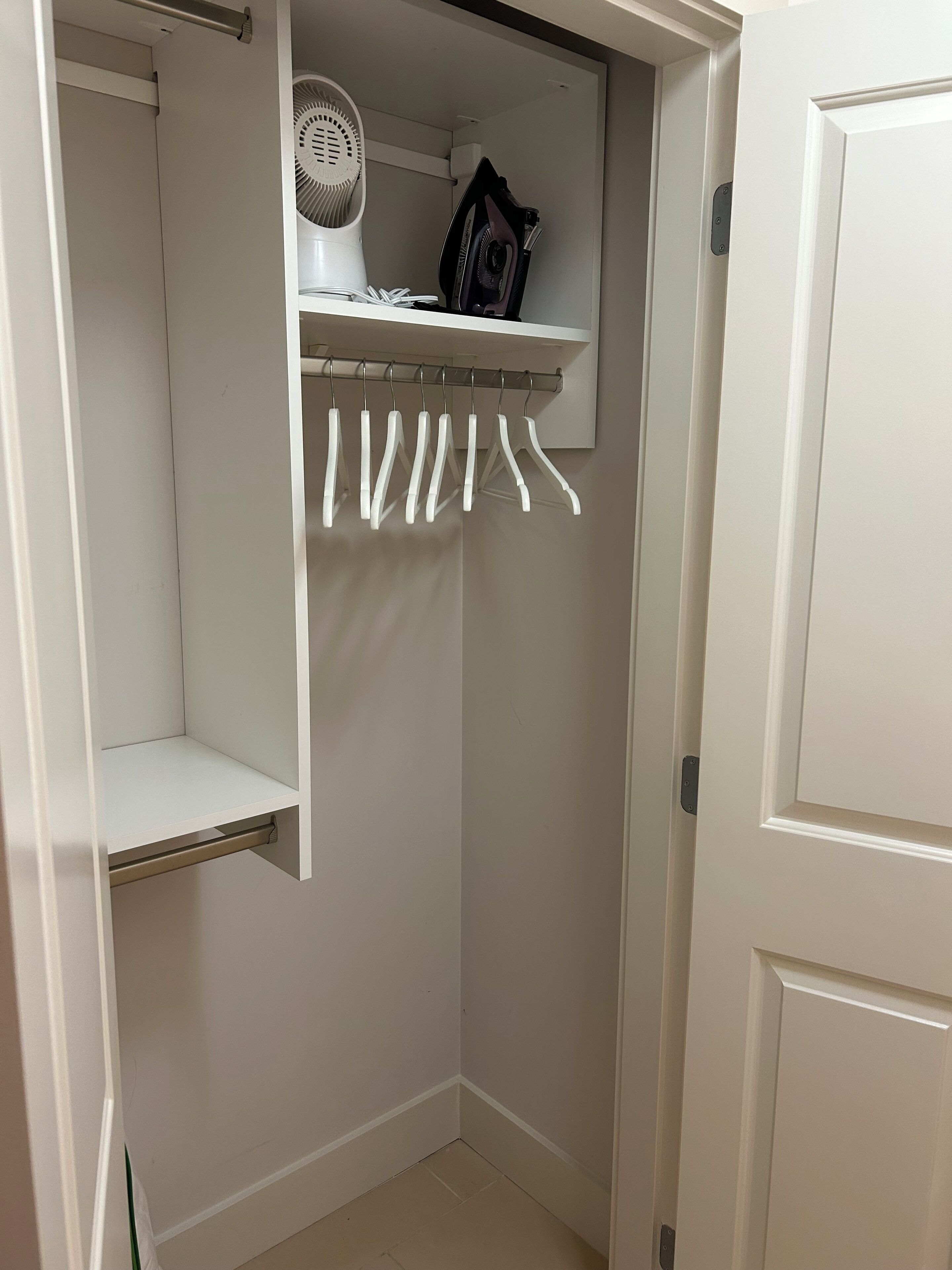 A white closet with a shelf holding a fan, an iron, and a few hangers.