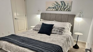 A neatly made bed with a grey headboard, two bedside lamps, a decorative leaf painting above the bed, and a small round table beside the bed. - Fern Hollow - unwind in this nature inspired suite in Edgemont Village. (North Vancouver)