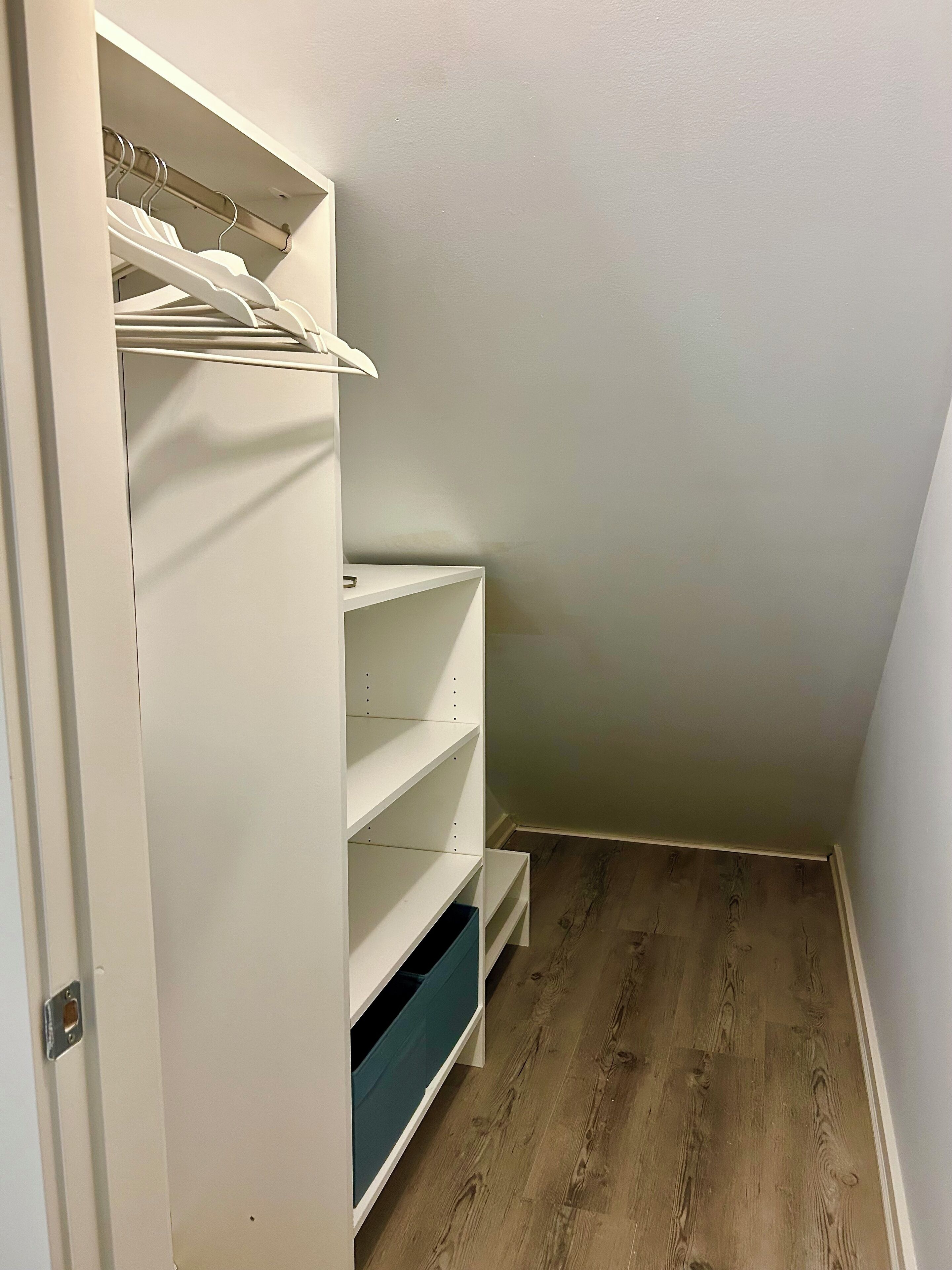 Un dressing accessible sans porte, doté d’étagères ouvertes, d’une tringle à vêtements et d’un bac de rangement bleu.