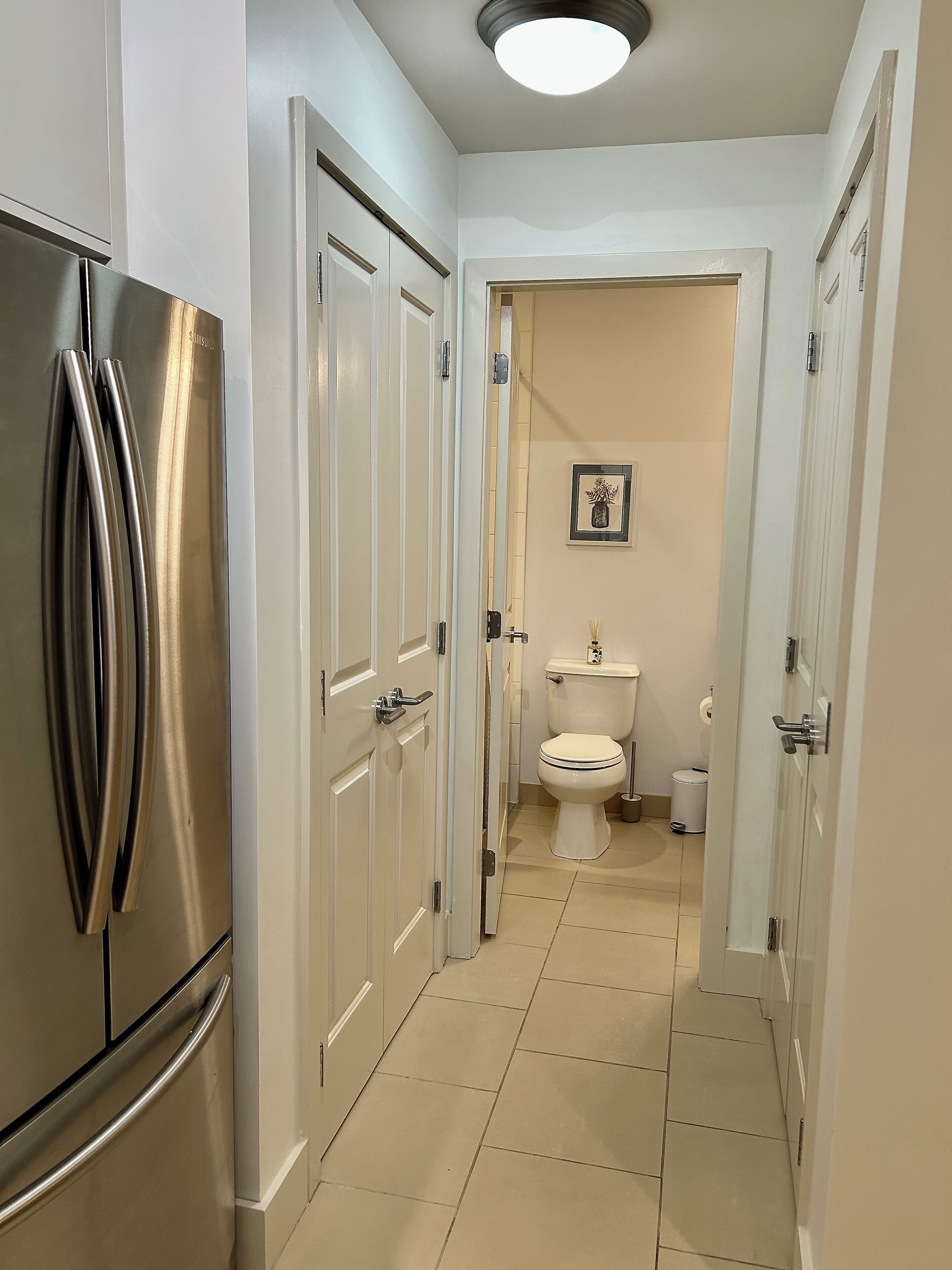 Ein schmaler Flur mit einem Edelstahl-Kühlschrank links, weißen Türen an beiden Seiten und einer Toilette, die durch eine geöffnete Tür im Hintergrund sichtbar ist.