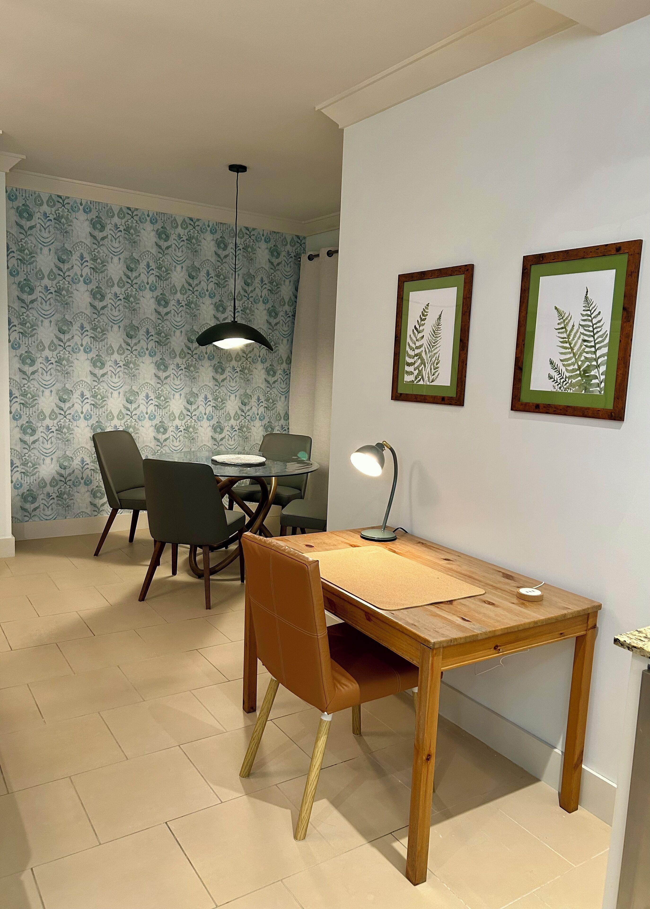 Un coin repas avec une table en bois, des chaises et une suspension. Deux reproductions botaniques encadrées ornent le mur.