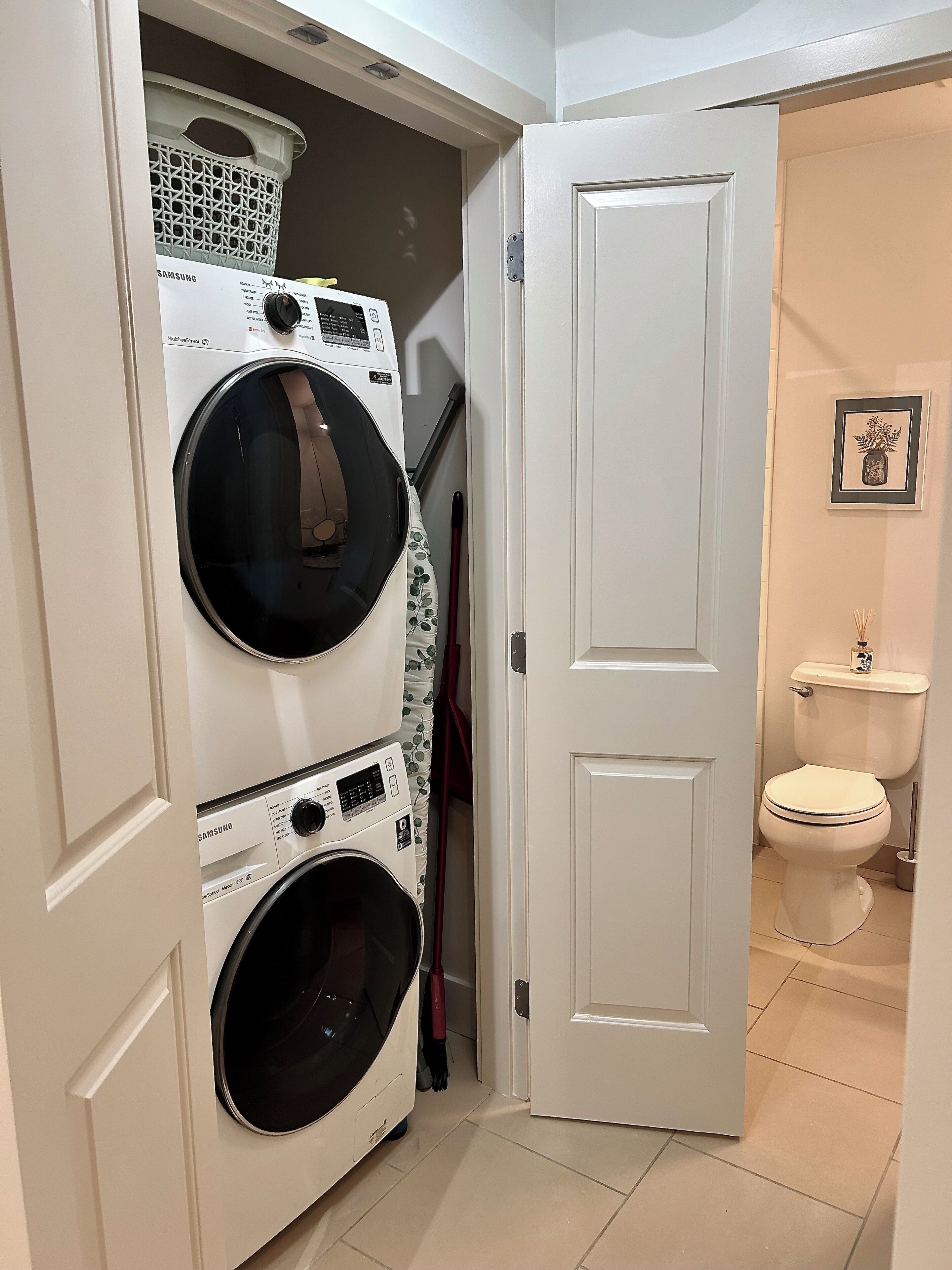 Une buanderie avec des machines à laver et des sèche-linge superposés, un balai et des toilettes visibles à travers une porte ouverte.