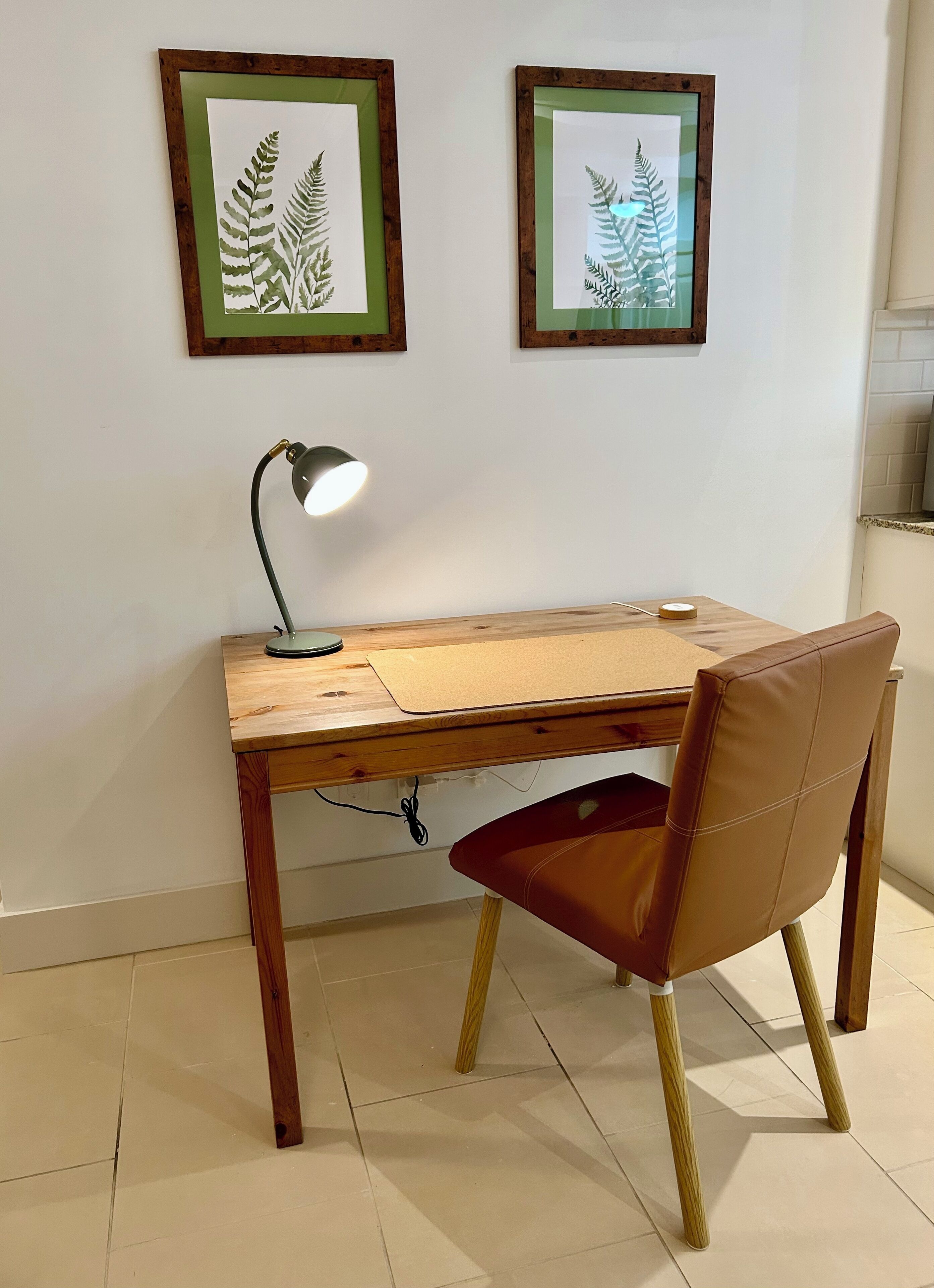 A wooden desk with a leather chair, a desk lamp, and two framed botanical prints on the wall.