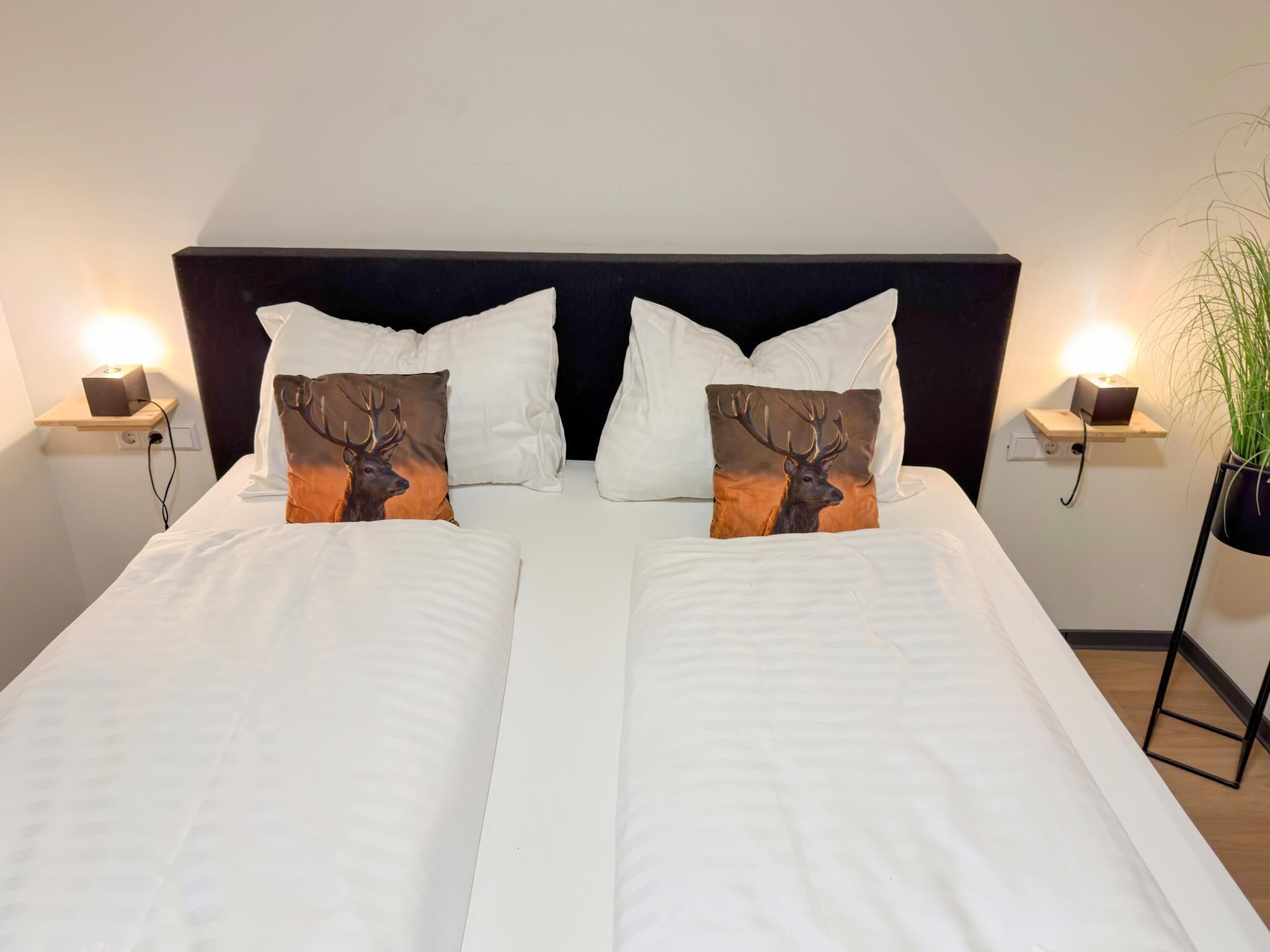 A double bed with white linens, two decorative pillows with deer images, a dark headboard, bedside tables with lamps, and a potted plant on the right.