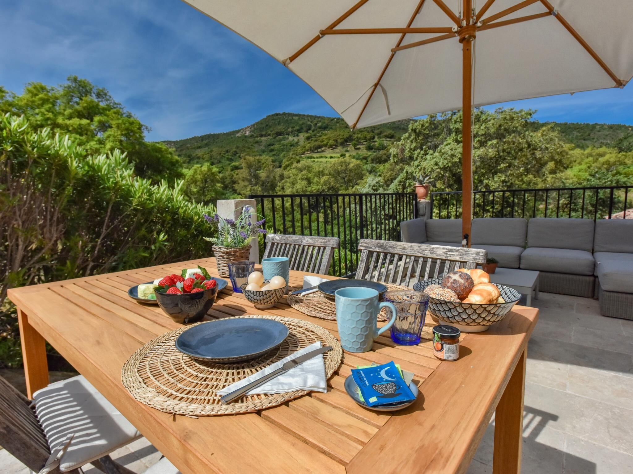 A wooden table with a woven placemat, a bowl of strawberries, a blue mug, and a basket of bread on an outdoor patio with a view of green hills.