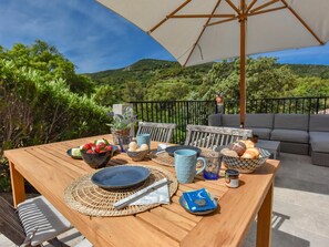 A wooden table with a woven placemat, a bowl of strawberries, a blue mug, and a basket of bread on an outdoor patio with a view of green hills. - Charming cottage on an intimate estate with pool (Le Plan de la Tour)