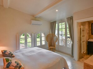 A bedroom with a large bed, a wicker chair, a view through a window, and a bathroom with a towel rack. - Charming cottage on an intimate estate with pool (Le Plan de la Tour)