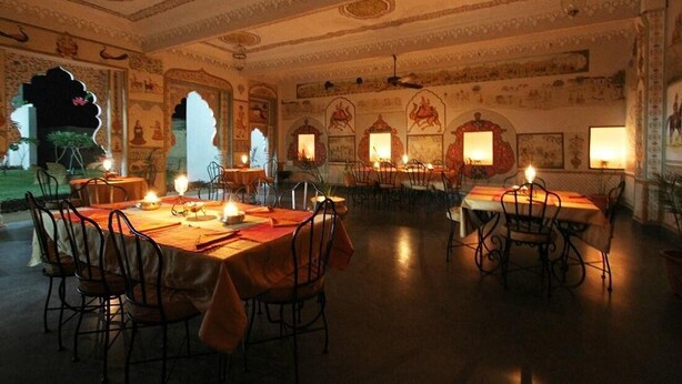 A traditional dining room with ornate ceiling and wall decorations, multiple tables set for dining, and candle lighting. - Barsingha Villa (Bap)