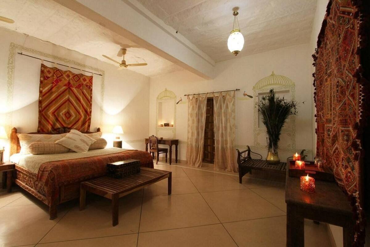 A spacious bedroom with a large bed, a wooden bench, a desk, and a chair. There are two lamps, a ceiling fan, and a decorative vase with candles.