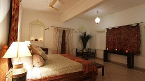 A bedroom with a bed, a nightstand, a lamp, a ceiling fan, a mirror, a window with curtains, a table with candles, and a decorative wall hanging. - Barsingha Villa (Bap)