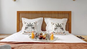 Een net opgemaakt bed met witte lakens, twee kussens met palmprentmotieven, een dienblad met drankjes en snacks, en een houten hoofdbord.