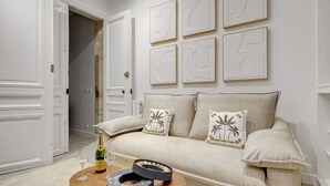 Een woonkamer met een beige bank, twee decoratieve kussens, een houten salontafel met een fles en glazen, en een wand met zes ingelijste kunstwerken.