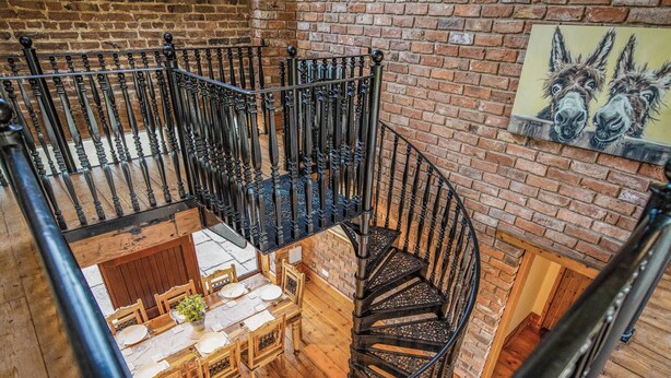 A spiral staircase with ornate black balusters, a dining area with wooden furniture, and a brick wall with a painting of donkeys. - The Threshing Barn - UKC8847 (Blankney Fen)