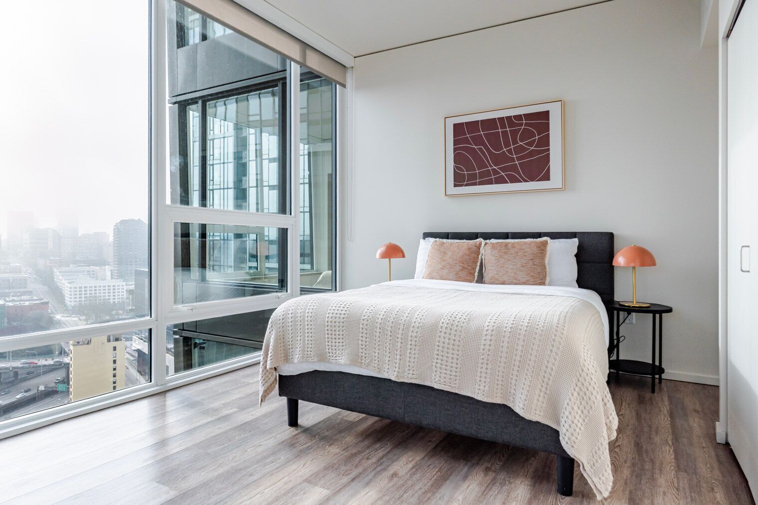A modern bedroom with a large window, a bed with a dark headboard, two bedside lamps, and a city view.