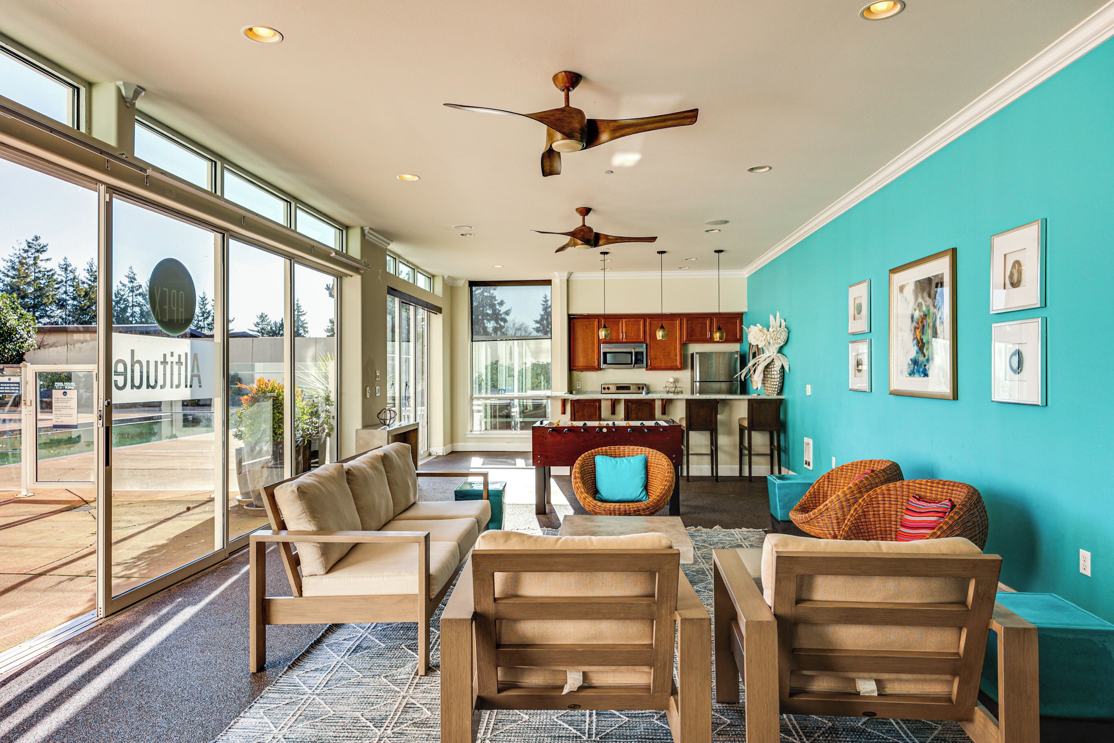 A spacious living area with a sofa set, a coffee table, and a dining table. There are ceiling fans, a kitchen area with a microwave, and a large window offering a view of the outdoors.