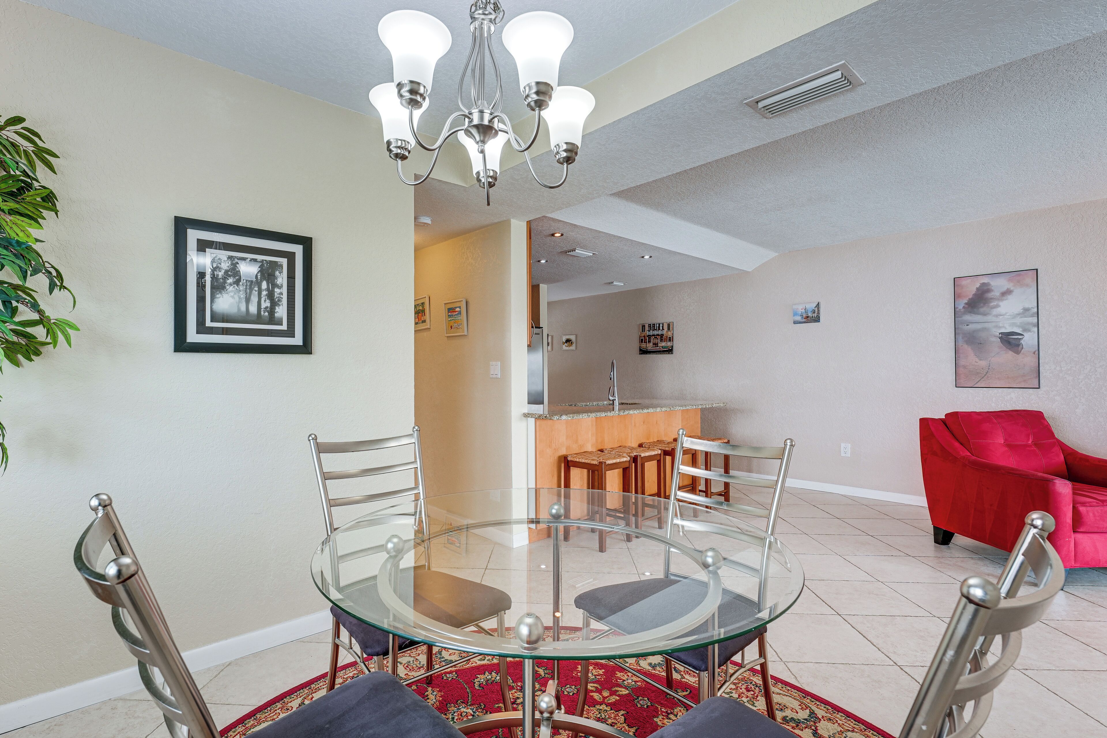 A dining area with a glass table and metal chairs, a red sofa, and a kitchen in the background.