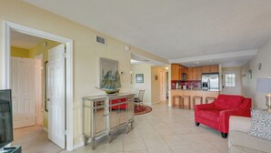 Un espacio habitable amplio con un sofá rojo, una mesa de centro de vidrio y un televisor. Se ve una cocina con armarios de madera y un refrigerador al fondo.