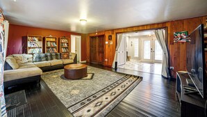 Ein geräumiges Wohnzimmer mit einer Couch, einem Hocker und einem großen Teppich. Bücherregale voller Bücher und ein Fernseher auf einem Holzgestell sind ebenfalls vorhanden.