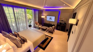 A spacious bedroom with a large bed, a TV, and a seating area with a view of greenery outside. - Kings Mansion (Candolim)