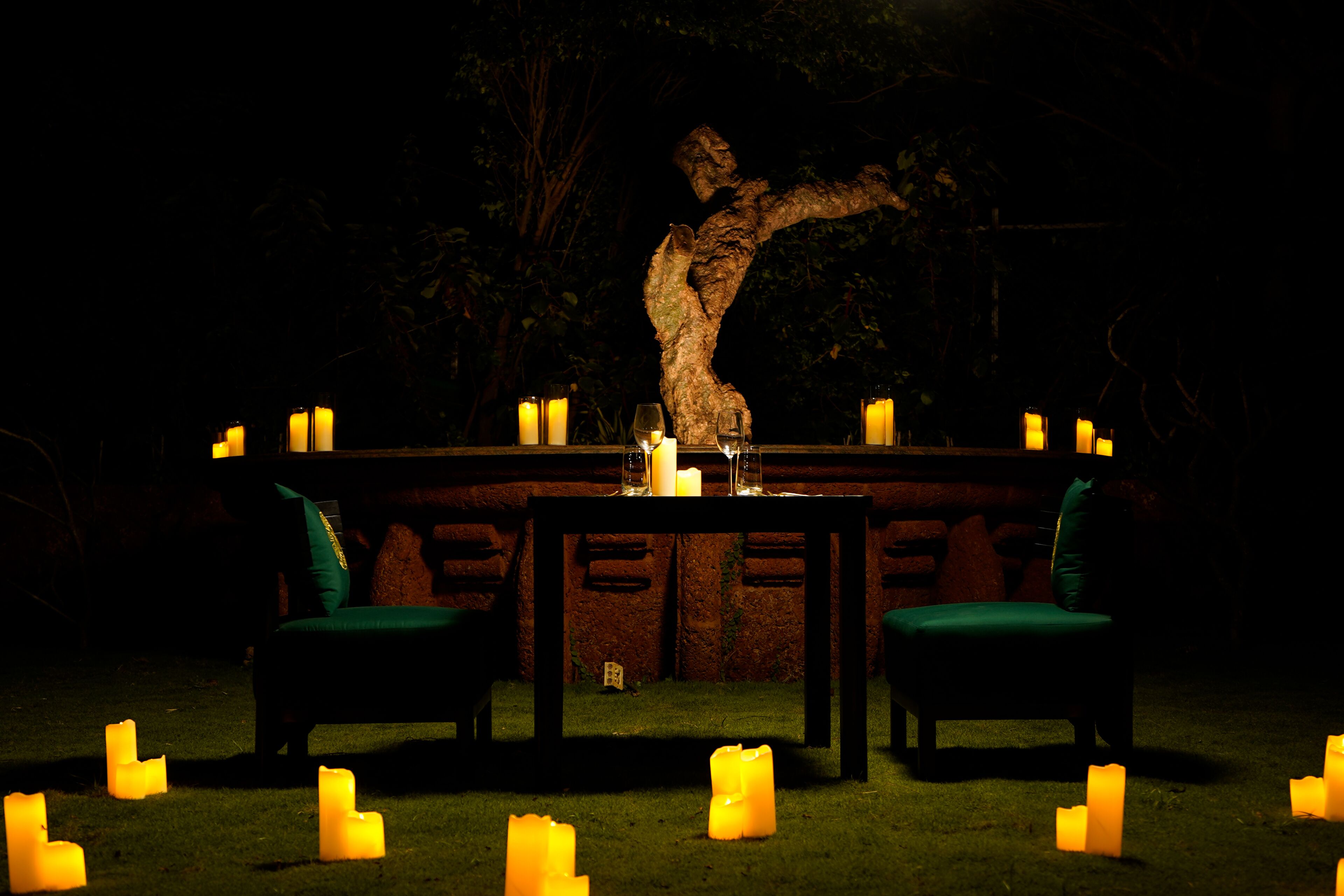 Un espacio exterior para comer con una mesa dispuesta para dos personas, rodeado de velas y un árbol al fondo.