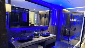 A modern bathroom with a large mirror, dark marble countertops, and blue ambient lighting. - Kings Mansion (Candolim)
