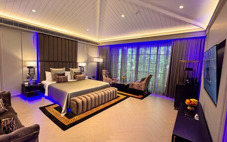 A modern bedroom with a large bed, a seating area, a TV, and a view of greenery outside.