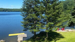Blick auf einen See mit Steg, einem großen Nadelbaum und roten Stühlen auf einer Wiese.
