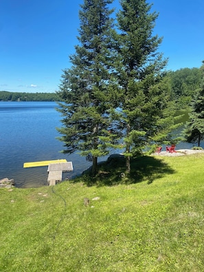 A lakeside view with a dock, a large evergreen tree, and red chairs on a grassy area. - Secluded Lakefront Cottage. (Haliburton)